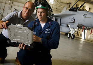  Picture of two men looking at laptop with F 18 in the background. U.S. Navy & Marine Corps Services