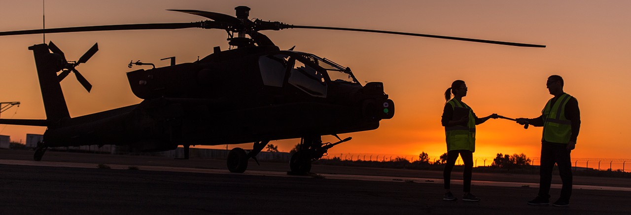 Two people standing on tarmac with Apache in background, with orange sunset backdrop.